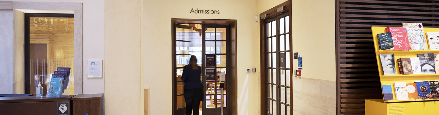 The door to the Admissions Office between an information desk and a shelf of books in Blackwell Hall at the Weston Library