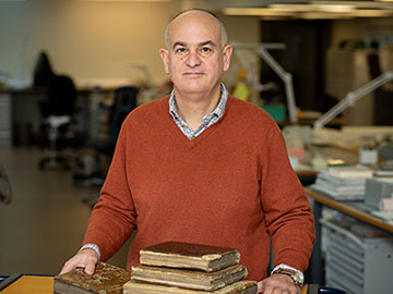 A man wearing an orange jumper and grey shirt stands in front of a table with old books