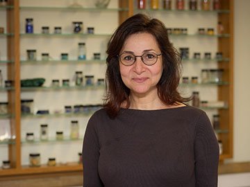 A woman with shoulder-length dark hair and glasses wearing a dark jumper stands in front of wooden shelving