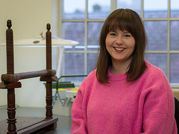 A woman with shoulder-length brown hair wearing a bright pink jumper stands in front of windows, next to a wooden frame