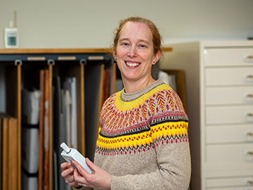 A woman wearing a grey, yellow and red Fairisle jumper stands in front of wooden storage and a filing cabinet