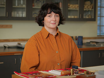 A woman with dark hair wearing an orange shirt stands in front of a table with materials