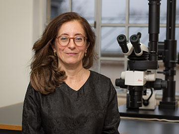A woman with long brown hair and glasses wearing a dark top stands in front of a microscope