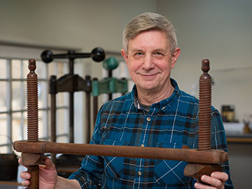 A man in a blue and green check shirt stands in front of a wooden frame