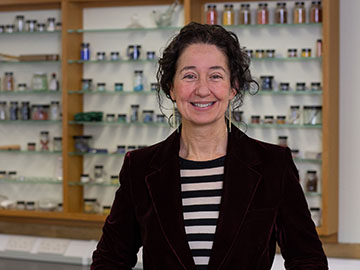 A woman with dark hair, wearing a dark suit jacket and striped top, stands in front of shelving
