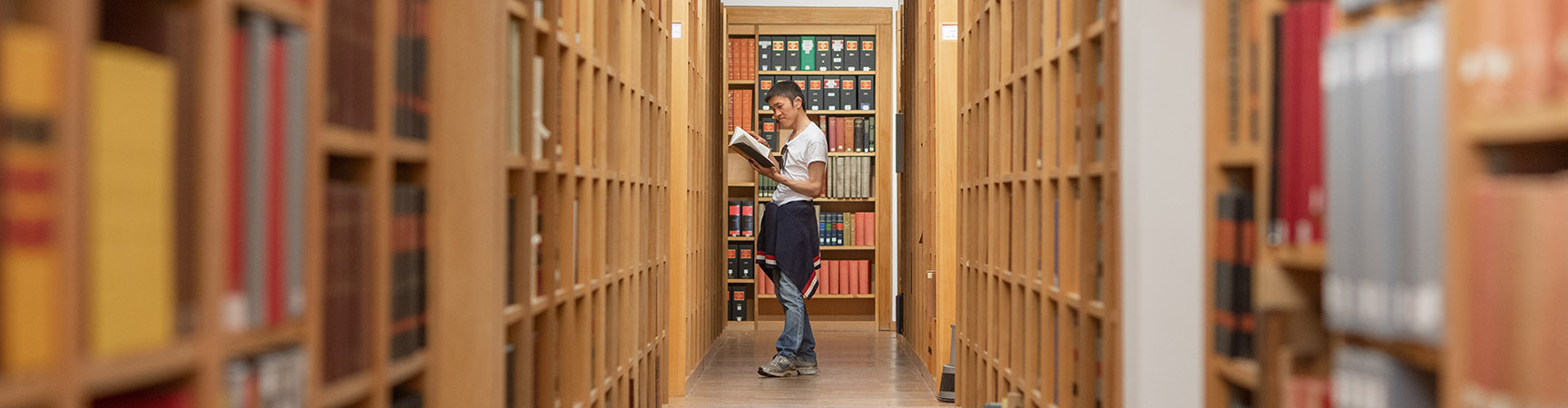 A long row of wooden bookshelves either side of a walkway, a student stands at the end reading