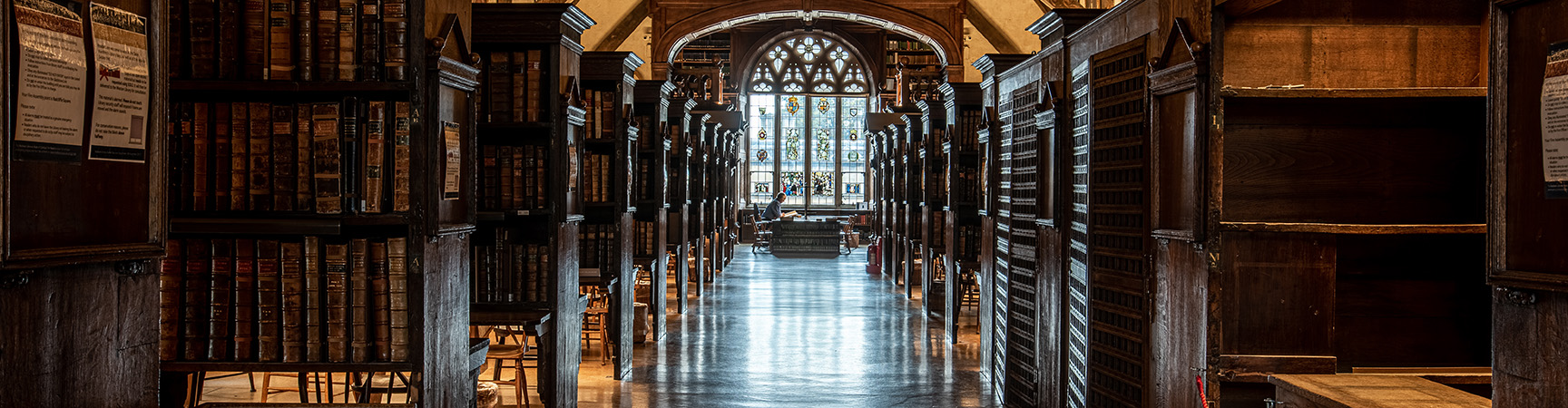 A long room with a stained glass window at the end, there are wooden book shelves and alcoves on either site