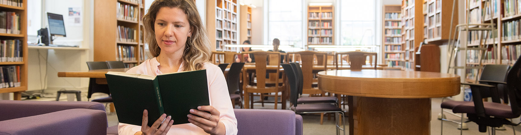 A student sits in a purple armchair reading a book, behind her is a library desk and there are bookshelves around the edge of the room