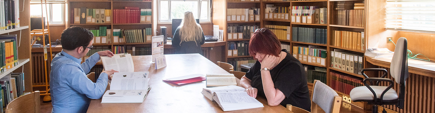 A large table in the middle of the room, two students sit either side each reading a book. There are bookshelves against the wall on either side