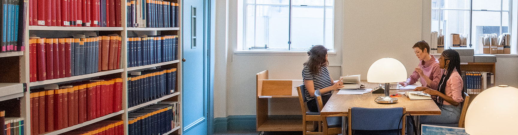 Three students sit around a wooden desk. They are in conversation. There is a bookshelf on one side of the room and large windows on another