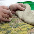 A detail - a pair of hands work on the tapestry - the back lining is held back and a small needle points towards the back of the threads of the tapestry