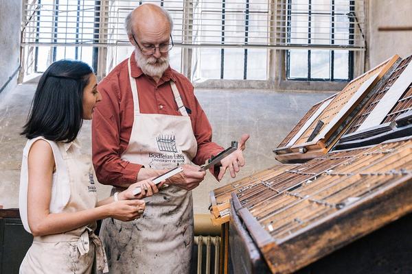 Two people in aprons stand beside a wooden display of printing blocks in front of a window