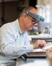 A side profile of a man - he has microscope glasses on his head - he holds a brush in his hand and works on a historic book