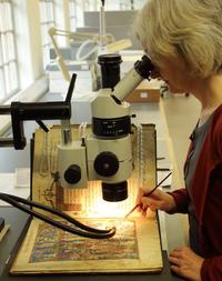 A woman looks through a microscope, beneath the microscope is an open book. The woman is using a paint brush to carry out conservation work on it.