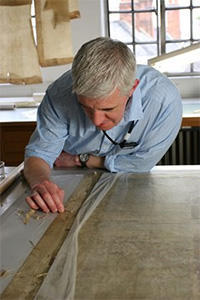 A man leans over a desk - he holds a small implement in his right hand and is carrying out a delicate conservation task on a flat manuscript