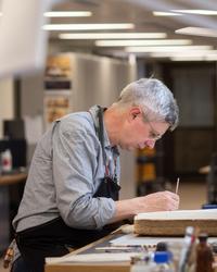 A side profile of a man - he is sitting down at a desk looking down, a book is open in front of him and he works on it with a small implement